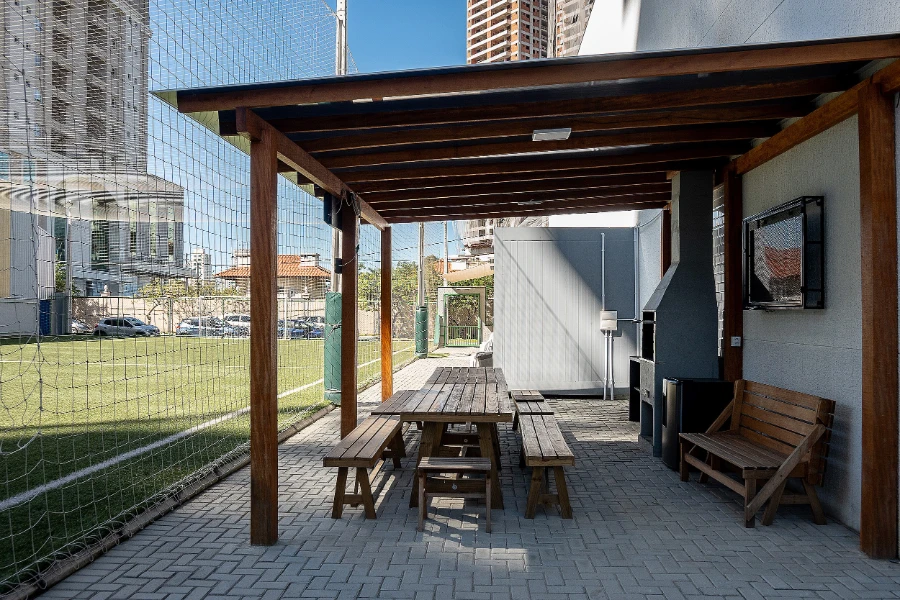 Área de convivência do Colégio Atlântico com mesas de madeira e cobertura de pergolado, localizada ao lado do campo esportivo.